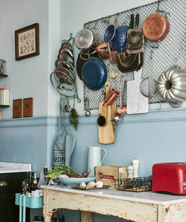 Pots, pans and cooking utensils hanging from large mesh grid on kitchen wall, over a rustic-looking side table.