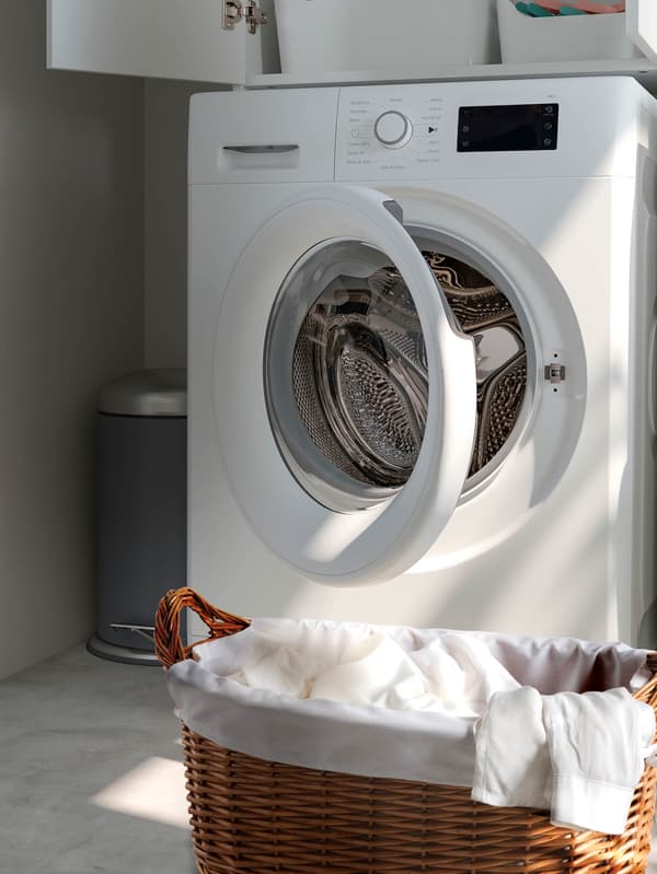Práčka UDDARP s pootvorenými dvierkami, pred ňou je prútený kôš na bielizeň TOLKNING.