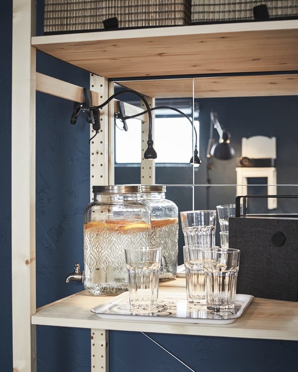 Prateleira da estante de madeira organizada como um tipo de minibar: recipientes de vidro com torneira cheios, tabuleiro com copos e coluna Bluetooth.