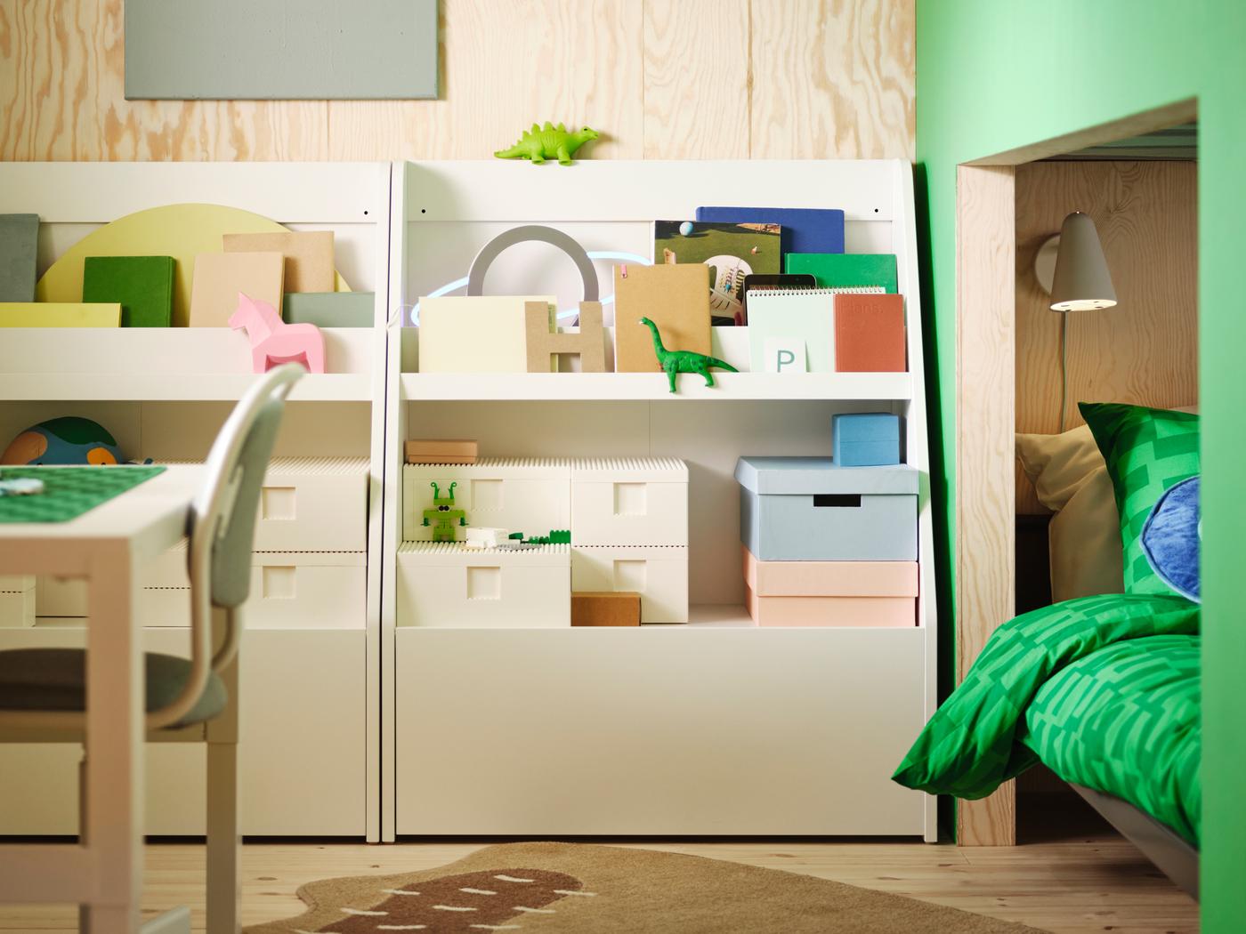 Pred leseno steno stojita dve beli BERGIG knjižni stojali s prostorom za shranjevanje, v katerih so različni zaboji, knjige in igrače.