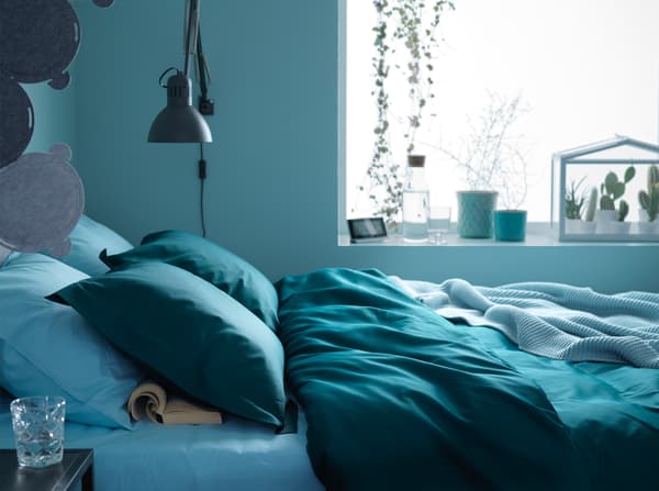 Près de la fenêtre d'une chambre bleue, lit garni de textiles en bleu et vert foncé.