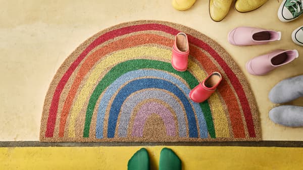 Preș de ușă PILLEMARK având forma și culorile curcubeului întins pe o podea galbenă pe care se mai află încălțăminte colorată și o pereche de picioare în șosete verzi.