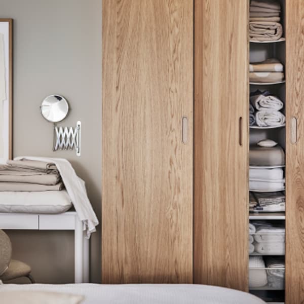 Près d’un lit, une armoire-penderie PAX/TONSTAD en blanc/chêne plaqué avec une porte ouverte révélant des textiles pliés rangés sur ses tablettes.