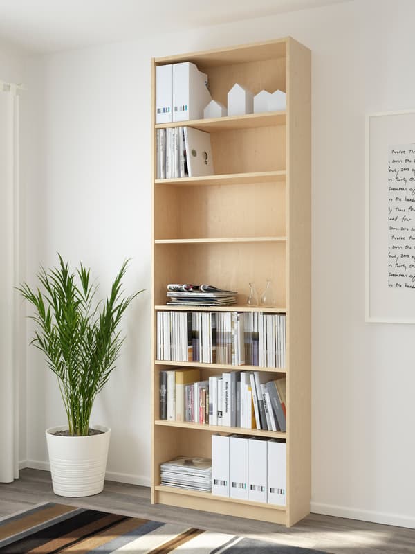 Prestatgeria alta de fusta clara amb baldes parcialment plenes de revistes, carpetes i objectes decoratius, al costat d’una planta en test i paret blanca amb quadre.