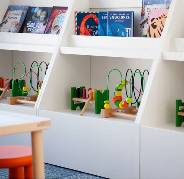 Prestatgeries infantils blanques amb llibres il·lustrats i jocs de fusta amb circuits de filferro i boles de colors, al costat d’una taula baixa.