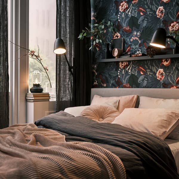 Pri tmavej stene s kvetinovým vzorom je posteľ, ktorá má po bokoch čierne stojacie lampy na čítanie SKURUP. V okne je zase stolová lampa TÄRNABY.