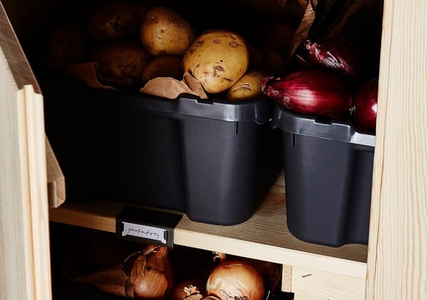 Primeiro plano de patacas e cebolas vermellas gardadas en cubos de plástico negro nun armario con estante de piñeiro.