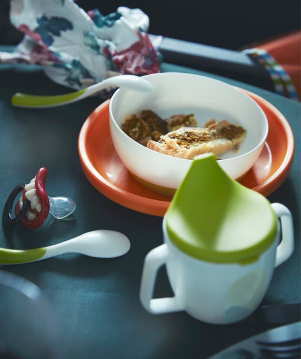 Primeiro plano dunha louza adaptada para bebé, incluído un vaso con tapa, culleres para alimentar o bebé e unha cunca.