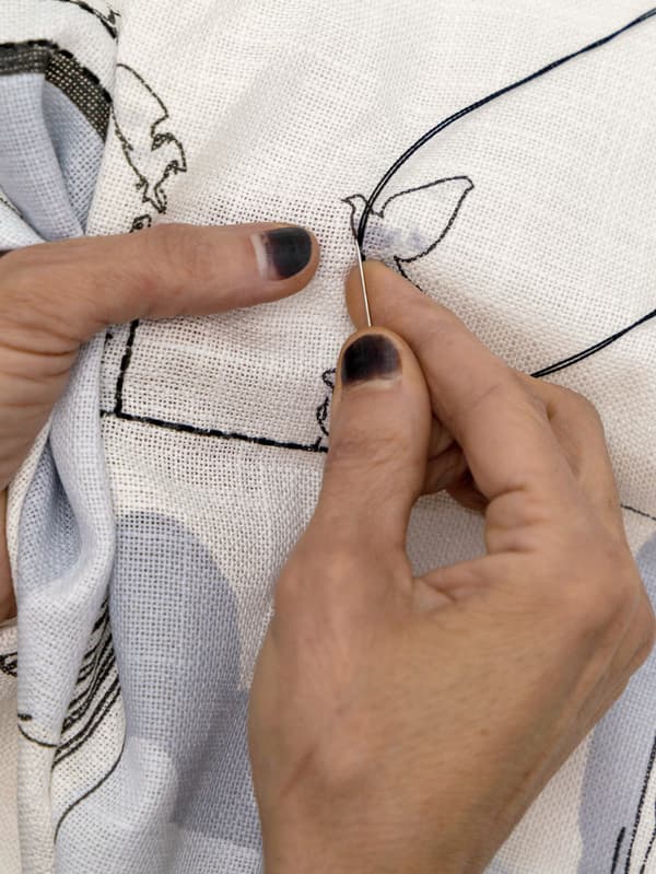 Primer plano de las manos de una artesana bordando una paloma y otros detalles en una funda de cojín.