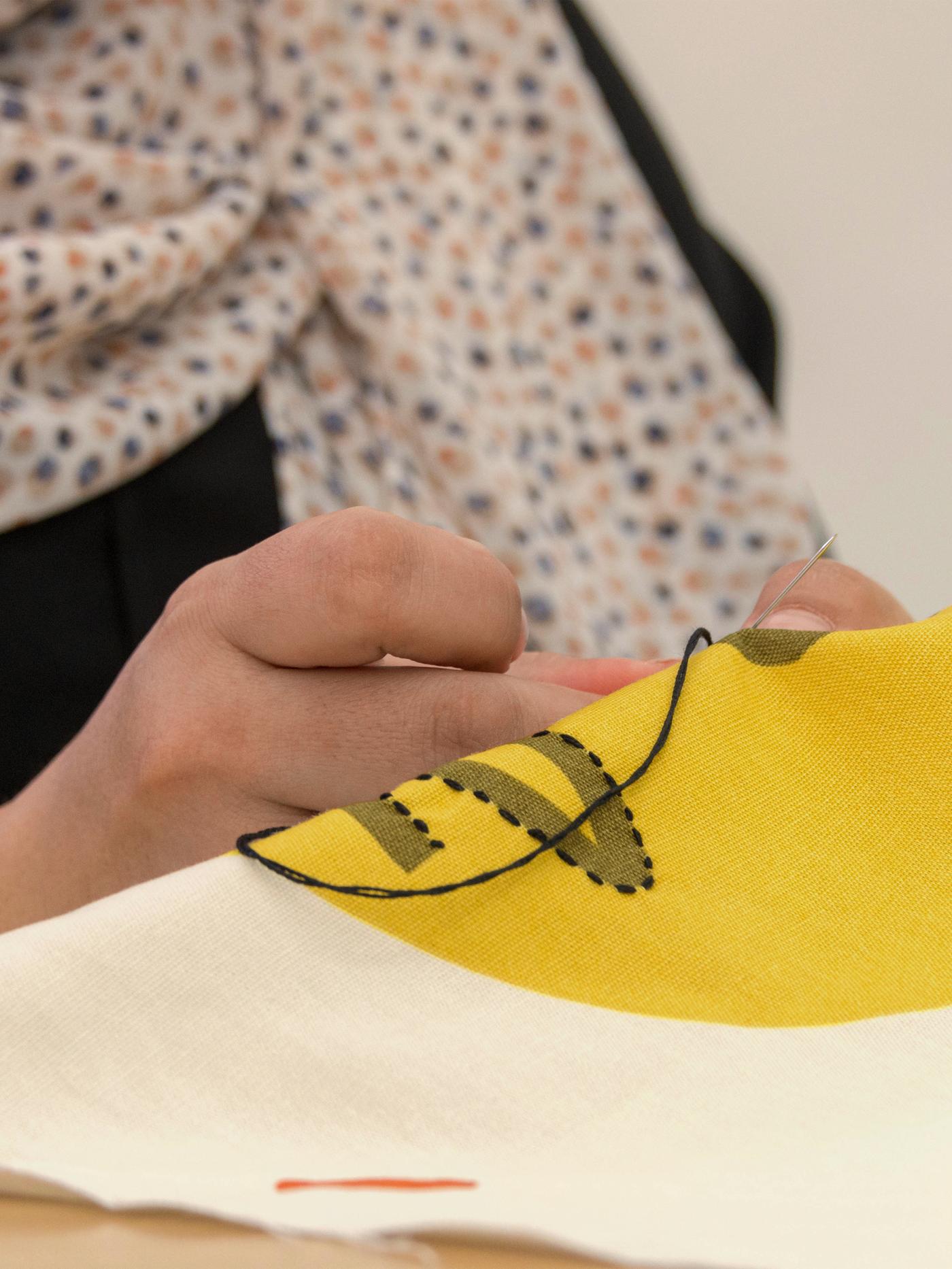 Primer plano de las manos de una artesana mientras borda la cara de una funda de cojín MANDELPIL.