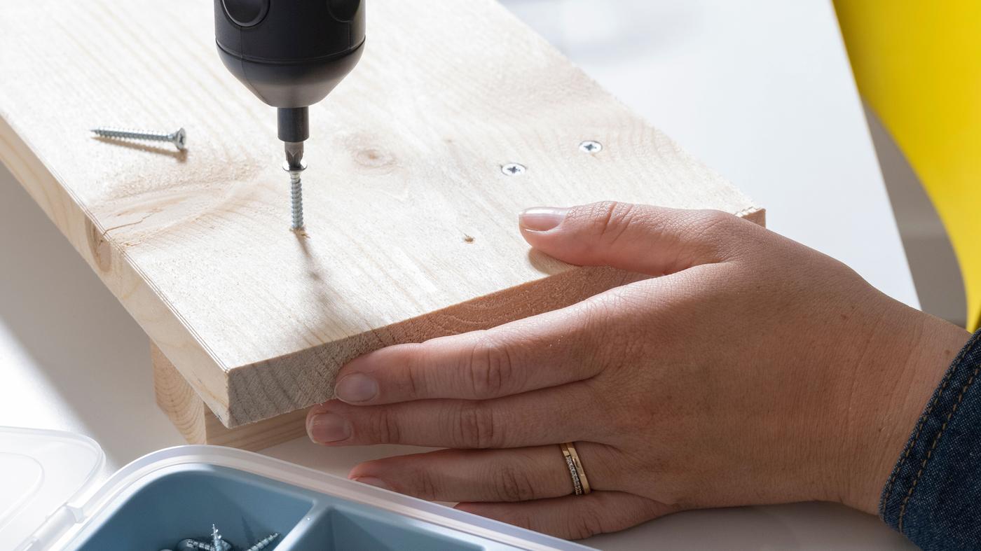 Primer plano de las manos de una mujer atornillando un tornillo en madera con un destornillador eléctrico.