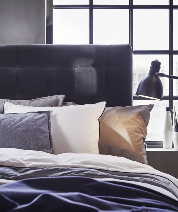 Primer plano de una cama con capas de ropa de cama y almohadas en blanco y gris, un cabecero oscuro y una lámpara de noche negra.