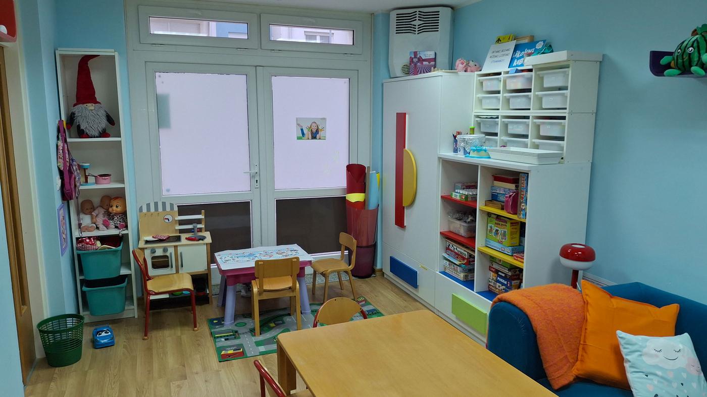 Prostorija uređena kao dječji kutak s plavim zidovima, drvenim podom i bijelim vratima s prozorima; sadrži mali stol s četiri stolice, police s igračkama i knjigama, ormar s plastičnim ladicama, kauč s narančastom dekicom i jastukom te šareni tepih.