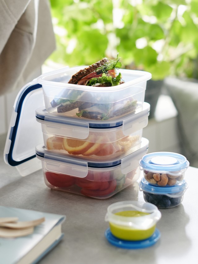 Průhledné plastové dózy IKEA 365+ s modrými víčky, naskládané na sobě, obsahují sendviče, plátky pomeranče a rajčata. Vedle jsou menší kulaté dózy s oříšky, borůvkami a zelenou omáčkou.