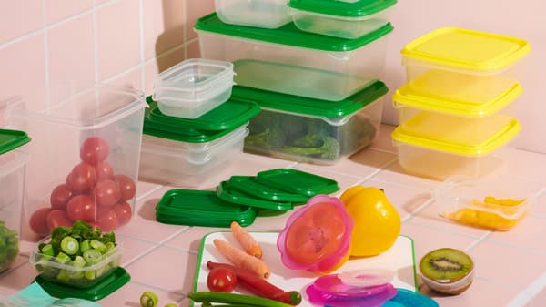 PRUTA food containers in various sizes, with yellow and green lids, are stacked neatly in the corner of a pink-tiled background. Fruits and vegetables are placed in front of the containers.