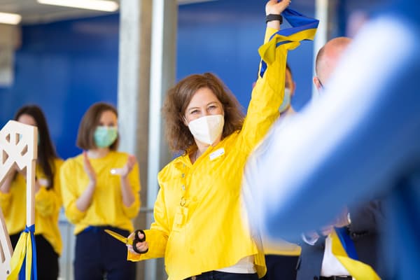 Przed sklepem IKEA, na środku stoi kobieta - pracowniczka sieci, która macha szwedzką flagą. Na drugim planie inni ludzie.