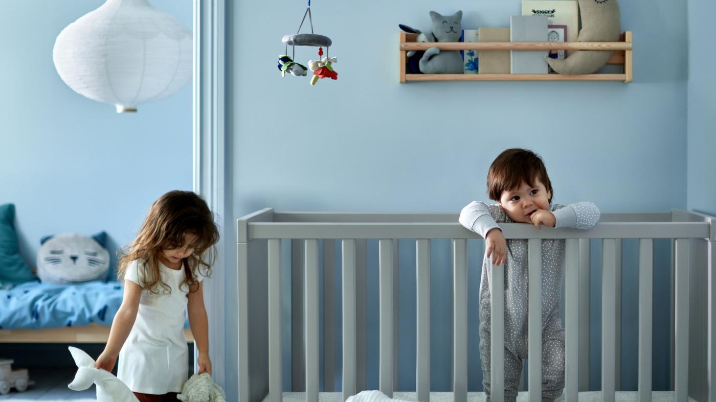 Puha játékokat kezében tartó kislány áll egy szürke kiságyban álló kisgyermek mellett, a kiságy kék ágyneművel van bevetve.