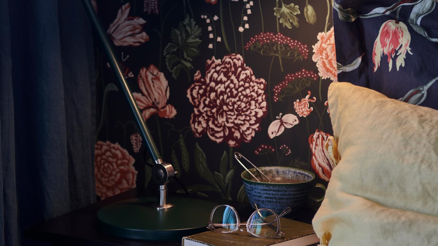 قاعدة مصباح عمل ‪ARÖD‬، وكوب وكتاب مع زوج نظارات على طاولة جانبية للسرير.