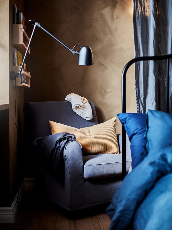 Quarto com poltrona GRÖNLID num canto, por trás da cama de casal. Por cima da poltrona, um candeeiro de secretária foi fixado numa prateleira montada na parede.