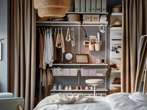 Quarto com um pequeno posto de trabalho com algumas prateleiras de um sistema de arrumação dentro de um roupeiro aberto com cortinados usados como portas.