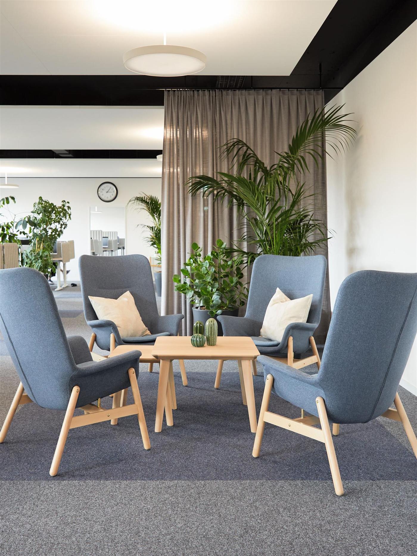 Quatre fauteuils gris autour d'une table en bois clair avec plantes, rideaux gris et horloge murale en arrière-plan.