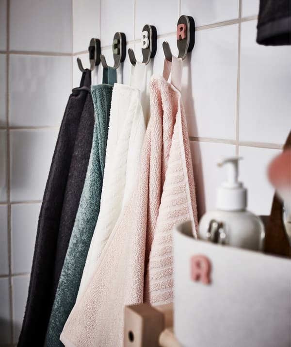 Quatre serviettes de différentes couleurs suspendues à quatre crochets dans une salle de bain en carreaux blancs.