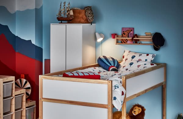 Racó d'una habitació infantil amb un llit reversible en la posició alta, un prestatge amb barana amb llibres i joguines, i un llum de llit encès.