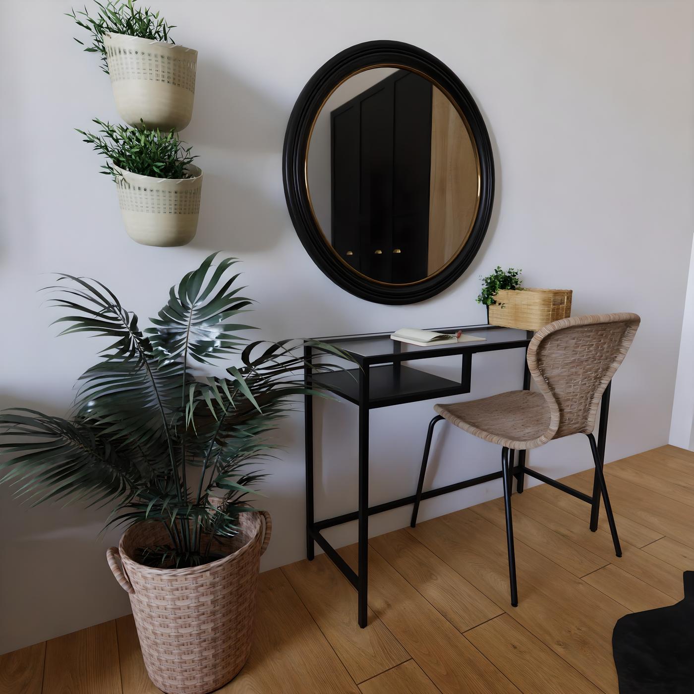 Radni stol s crnom metalnom konstrukcijom, okruglo ogledalo iznad, dvije viseće posude s biljkama i velika biljka u košari pored stola.