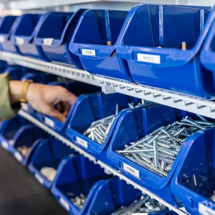 Raft cu cutii albastre pline cu șuruburi și piese de fixare, din zona de accesorii IKEA, unde o persoană își alege elementele necesare pentru montaj sau reparații.