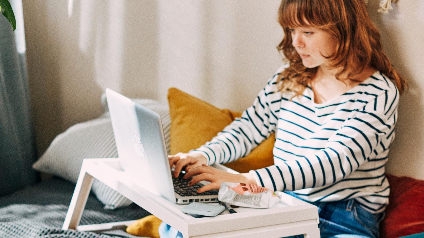 Ragazza dai capelli rossi, con maglione a righe, seduta sul letto mentre scrive su un computer portatile appoggiato su un vassoio da letto KLIPSK bianco.