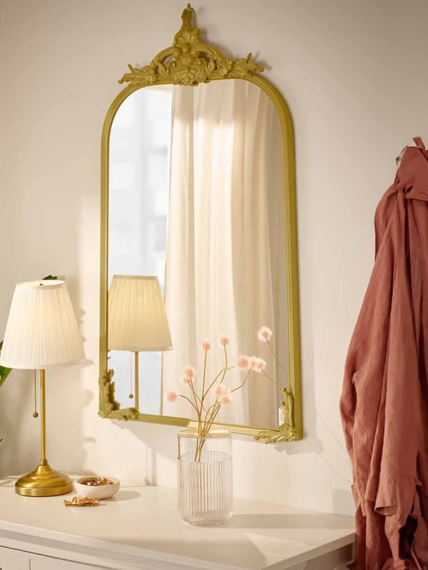RAMEBO gold mirror hanging on a white wall with a vase of flowers and a bedside lamp.