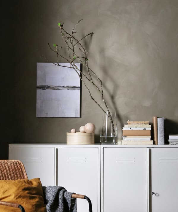 Rangée d’armoires blanches contre un mur gris. Au-dessus se trouvent des livres, des objets de décoration, un vase avec une longue branche et quelques bourgeons verts.