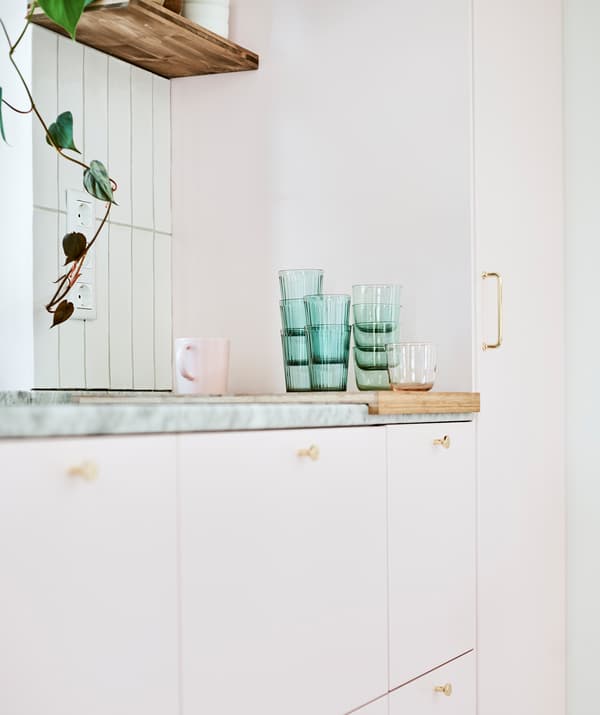 Rangée d’armoires de cuisine aux portes roses avec une pile de verres verts et une tasse rose sur une planche à découper posée sur un plan de travail en marbre.