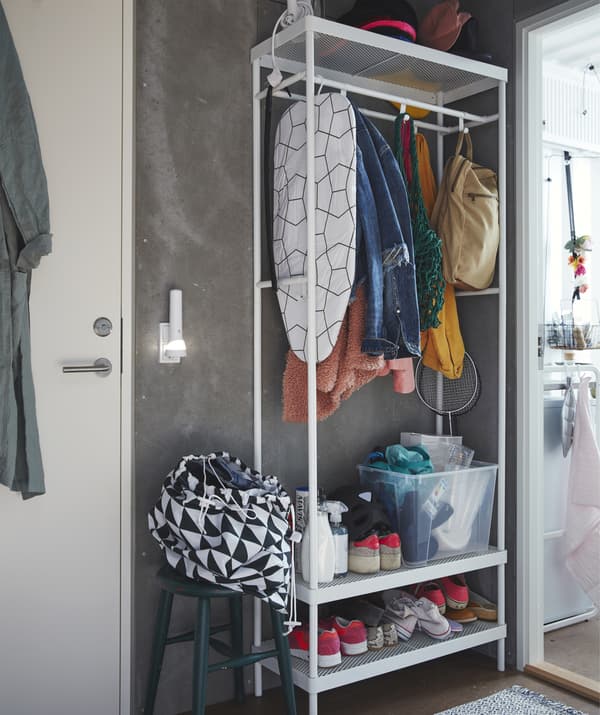 Rangement ouvert haut avec chaussures, manteaux et planche à repasser sur des tablettes et des barres, à côté d’une porte.