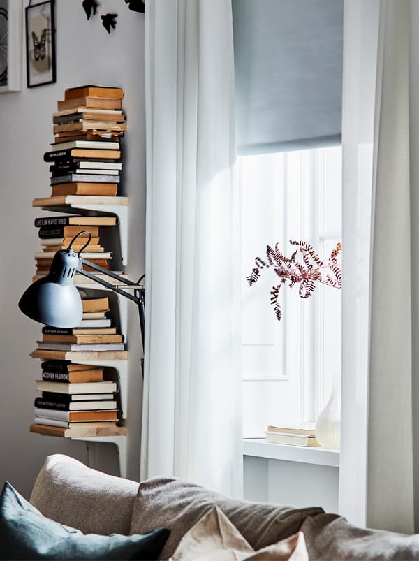 Rangement vertical de livres réalisé à partir de quatre consoles SIBBHULT et des étagères à côté d’une fenêtre habillée de voilages blancs.