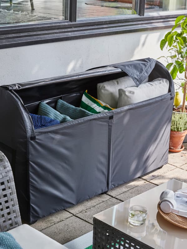 Rangements d’extérieur en gris foncé contenant de grands et de petits coussins, sur une terrasse, à côté de plantes en pots.