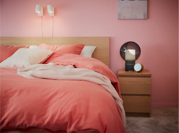 Rangka katil dan meja sisi katil dalam kesan kayu oak berwarna putih, tekstil katil berwarna coklat merah lembut dan lampu dinding berwarna putih.