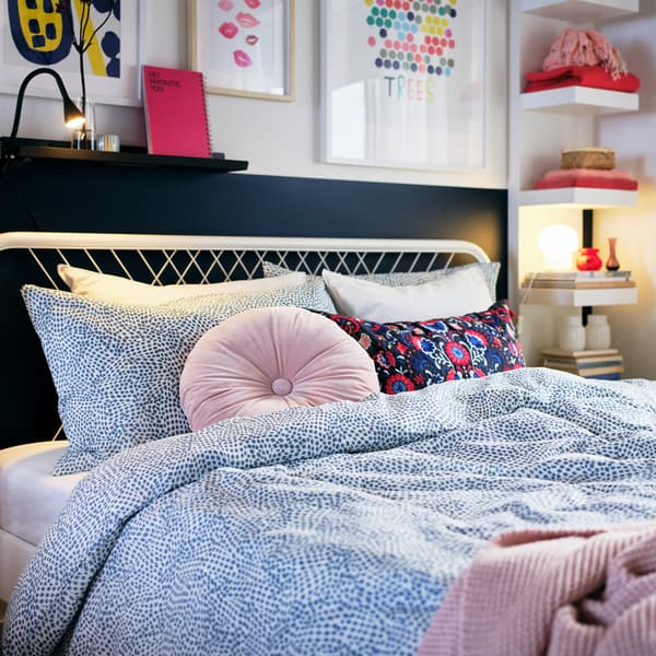 Rangka katil NESTTUN berwarna putih dengan duvet dan alas berwarna merah jambu dan kusyen bulat dengan karya seni dan para-para terbuka pada dinding di belakang.