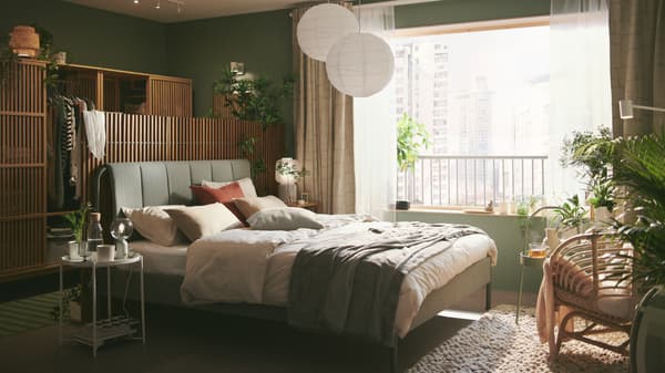 Rangka katil upholsteri TÄLLÅSEN berwarna kelabu-hijau dengan skrin privasi di belakangnya terletak di dalam bilik tidur berwarna hijau yang tenang dengan tingkap besar.