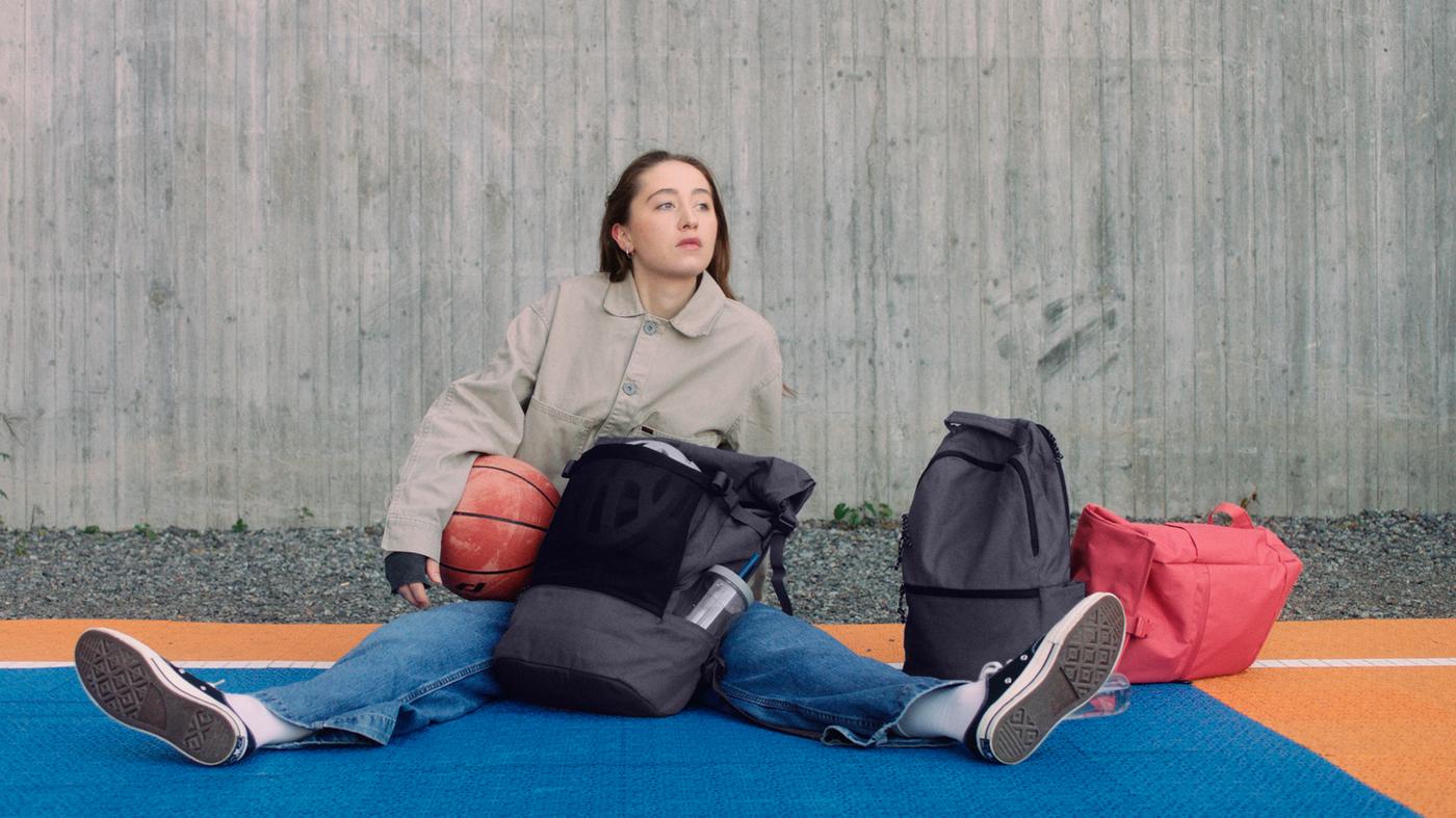 Rapaza suxeitando un balón de baloncesto sentada nunha pista de baloncesto, con bolsas DRÖMSÄCK, VÄRLDENS e STARTTID ao seu carón.