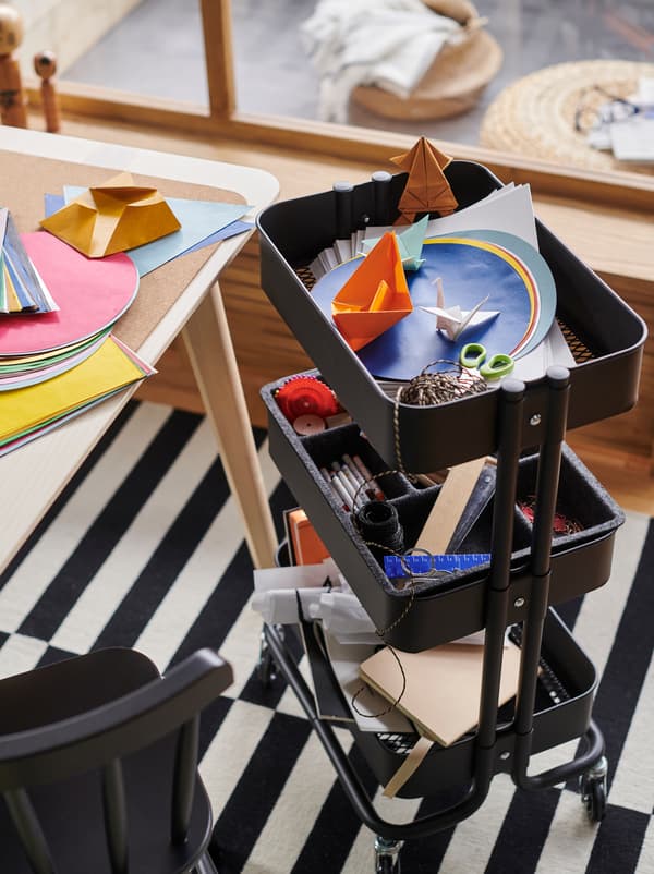 รถเข็น RÅSKOG/รวสกู๊ก สีดำใส่อุปกรณ์สำหรับทำงานศิลปะและงานฝีมือตั้งอยู่ถัดจากท็อปโต๊ะที่มีกระดาษสีวางอยู่อย่างกระจัดกระจาย