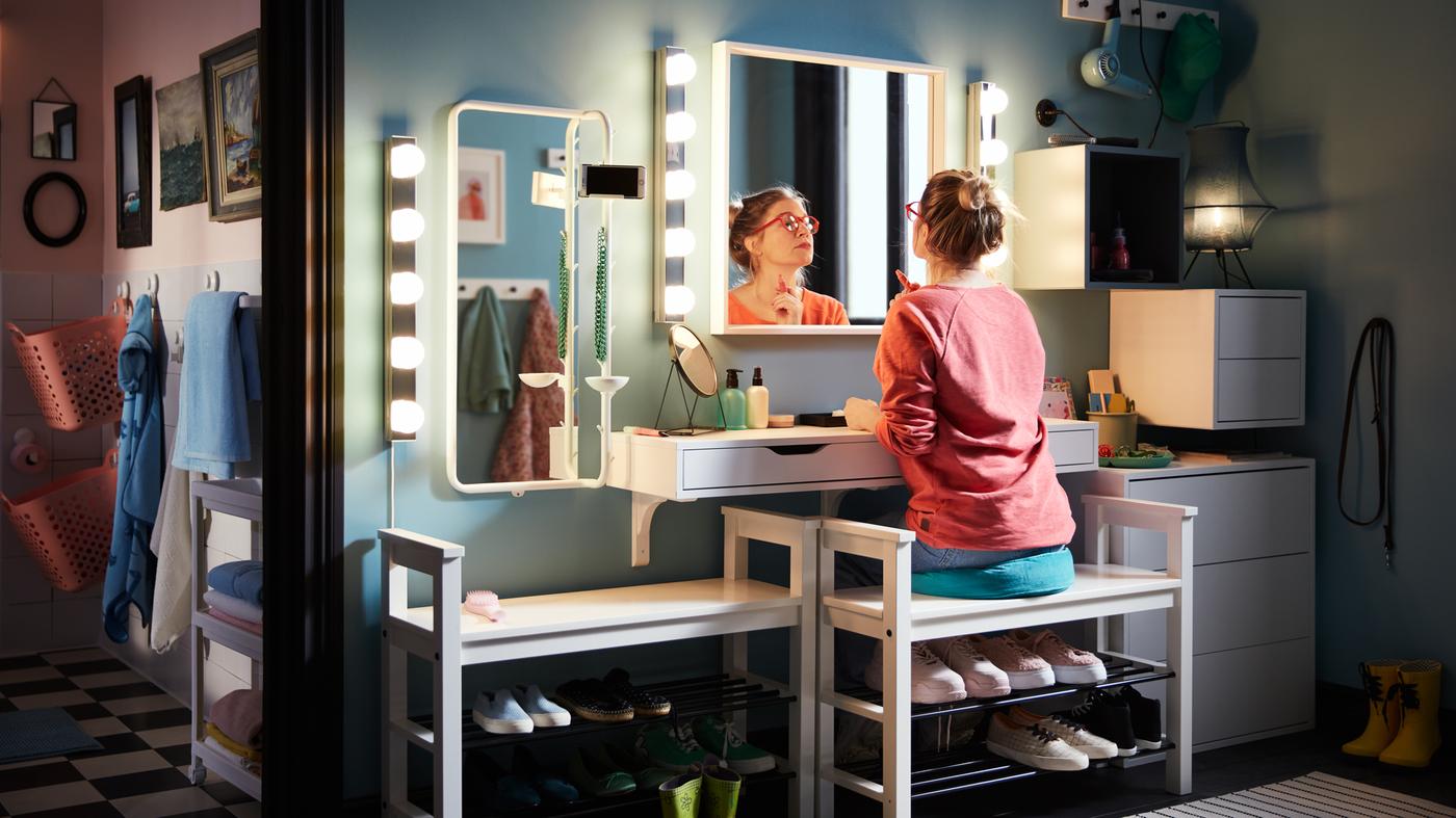 ردهة منزل حُوِّلت إلى مساحة للتزين باستخدام رف حائط ومصابيح إضاءة ومرايا، تجلس فيها امرأة على إحدى منصّات تخزين الأحذية وتضع مستحضرات التجميل الخاصة بها.