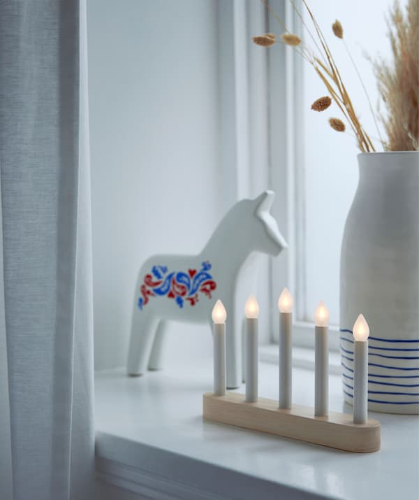 Rebord de fenêtre décoré avec un cheval de Dalécarlie, un candélabre LED et un vase en céramique contenant des tiges d’herbes séchées.