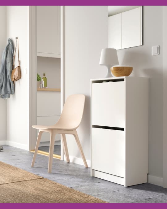 Recibidor con mueble zapatero blanco, silla clara y espejo sobre la pared, junto a un pasillo luminoso.