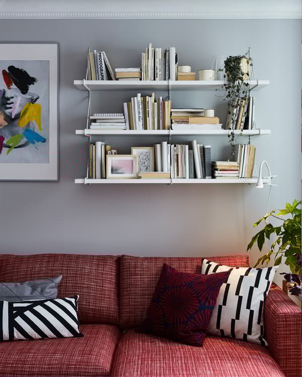 Red sofa with white and red patterned decorative cushions. White shelves are mounted on the wall behind and store books.