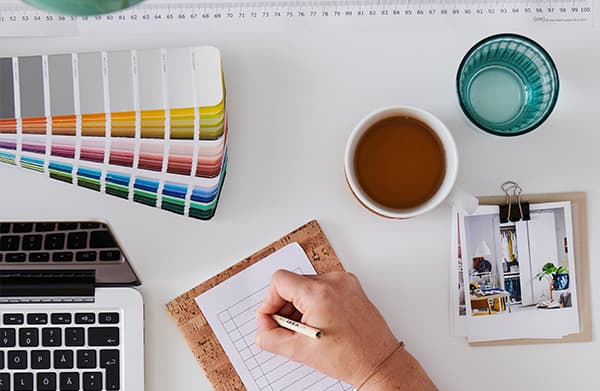 Register your interest for free message with an image. A white desk with a laptop, cup of coffee, glass of water, a pencil organiser cup, a desk light and a customer writing notes on the desk to get ready for an online planing service from home. 