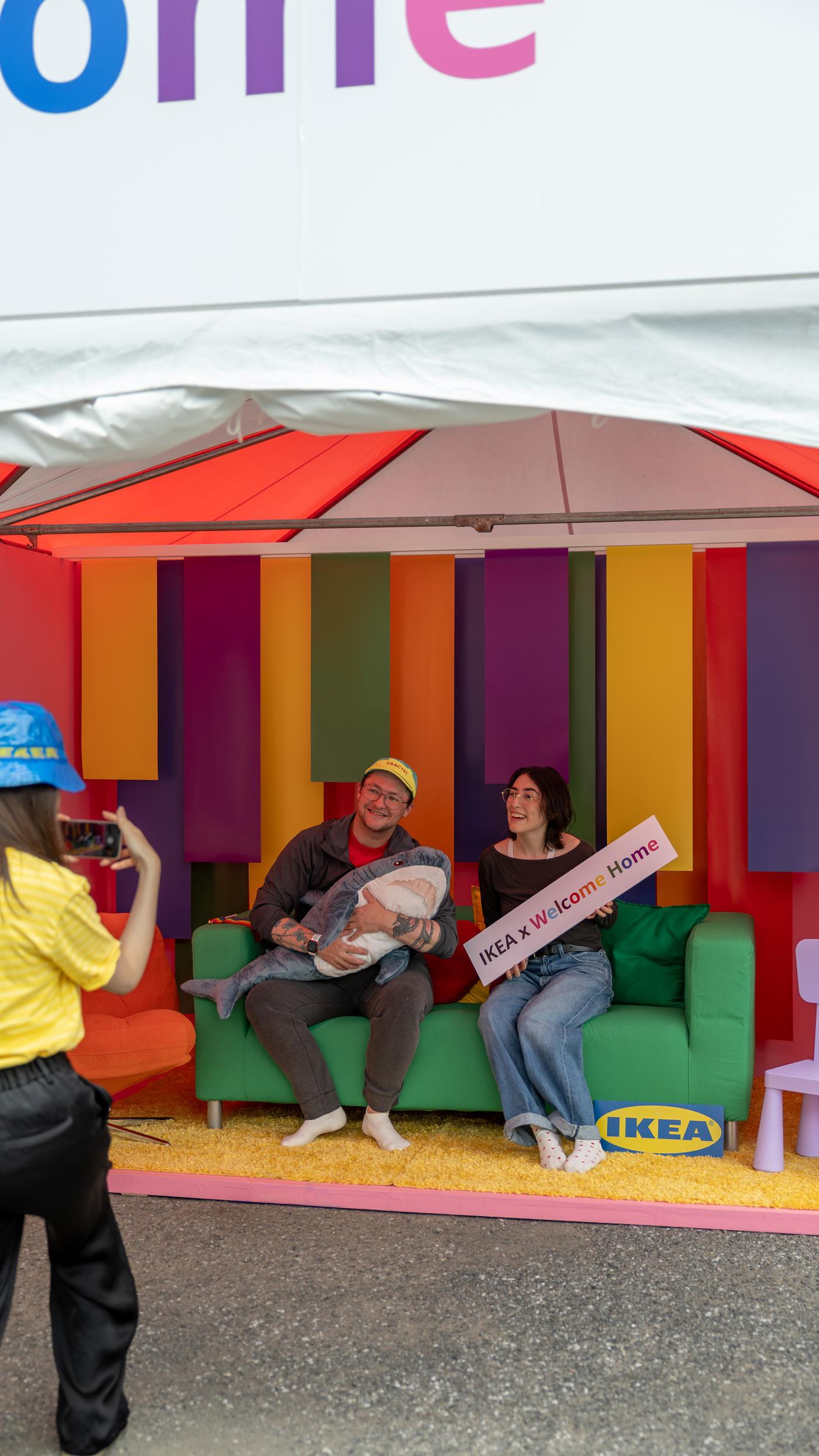 レインボーカラーの壁と緑とオレンジのソファが並ぶWelcome Homeブースで、家族がぬいぐるみとIKEA x Welcome Homeと書かれたサインを持ち、コワーカーが撮影している。