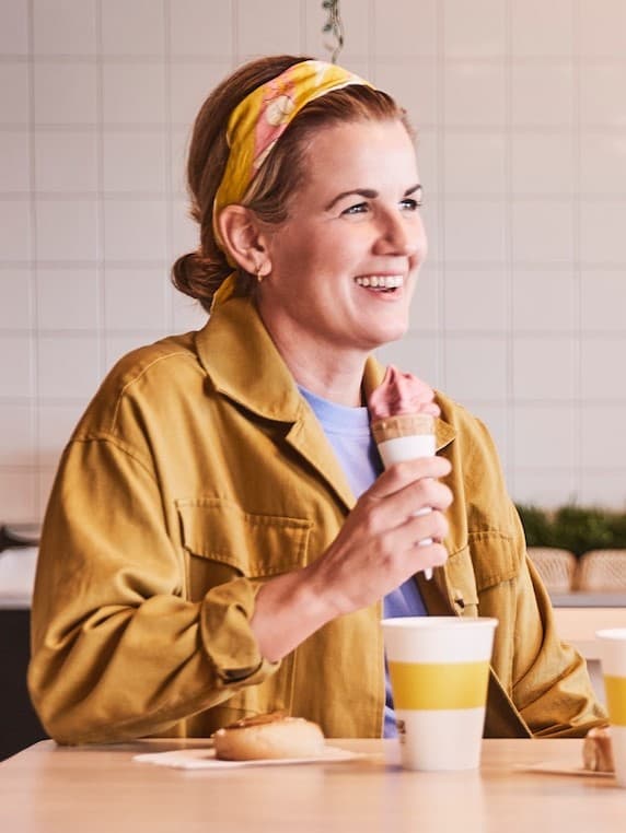 Ręka osoby ubranej w szarą bluzkę, która ma pomalowane na fioletowo paznokcie, trzyma truskawkowe lody w rożku.