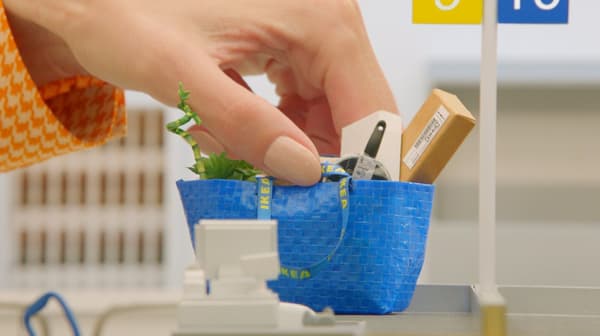 Ręka trzymająca miniaturową niebieską torbę IKEA, w której są miniaturowe produkty sieci.