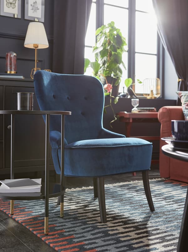 ห้องนั่งเล่นผนังสีเข้มที่มีแสงแดดส่อง มีอาร์มแชร์ REMSTA/เร็มสตา สีน้ำเงินตั้งอยู่ตรงมุมที่มีพรม RESENSTAD ที่มีลวดลายหลากสี
